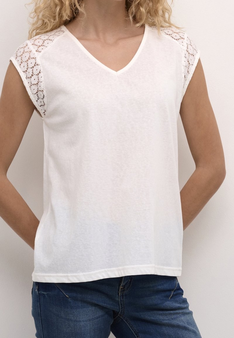 Femme portant un haut blanc à col en V avec des manches courtes en dentelle et un jean bleu, debout les mains derrière le dos.