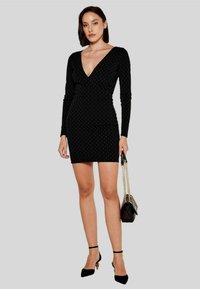 Vestito mini nero elasticizzato con maniche lunghe e scollatura a V profonda, caratterizzato da un motivo testurizzato. Abbinato a scarpe con cinturino alla caviglia nere e una borsa.