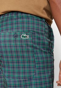 Zaļas svītrainas šortos ir aizmugurējā kabata ar pogu un Lacoste krokodila logo. Audumam ir gluda tekstūra un strukturēts dizains.