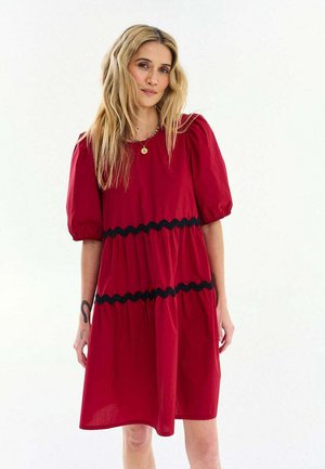 Kobieta o długich blond włosach, ubrana w czerwoną sukienkę do kolan z bufkami na rękawach i czarnymi falistymi taśmami, stojąca na białym tle.