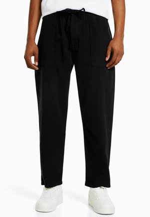 Mężczyzna ubrany w luźne czarne spodnie z przednimi kieszeniami, białe trampki i białą koszulę, stojący na jednolitym tle.