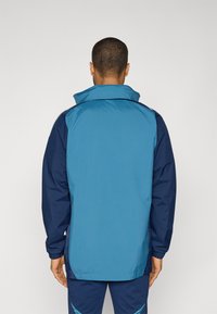 Blauwe en marineblauwe windbreakerjas met een hoge kraag, elastische manchetten en een ontspannen pasvorm. Gladde textuur en lichtgewicht stof.