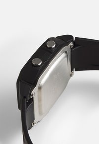 Montre numérique noire avec un boîtier carré en acier inoxydable, deux boutons ronds sur le côté et un bracelet souple et flexible noir.