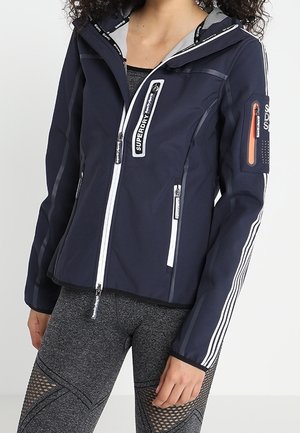 Frau trägt eine marineblaue Superdry-Zipjacke mit weißen Streifen an den Ärmeln und graue Leggings mit Netzpaneelen.