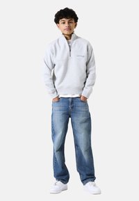 Sudadera gris con cremallera y cuello alto, con el texto azul "ÚNETE AL CLUB", combinada con jeans azules holgados y zapatillas blancas.