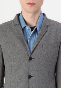 Grauer Blazer mit einem Hahnentrittmuster, zwei Knöpfen und einer Brusttasche, über einem blauen Denimhemd mit Kragen und Knopfleiste getragen.