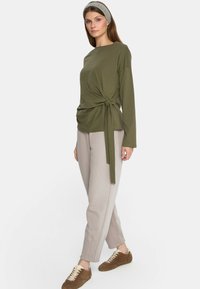 Oliwkowy top z długim rękawem i wiązanym przodem, w zestawieniu z jasnoszarymi spodniami i brązowymi sneakersami. Gładki materiał i luźny krój.