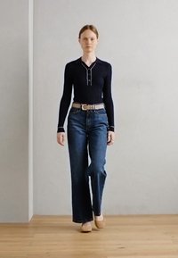 Dark blue ribbed long-sleeve top with a small collar, paired with high-waisted wide-leg jeans and light brown shoes. Minimalist backdrop.