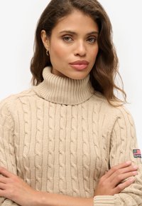 Beige kabelgebreide coltrui met geribbelde textuur, voorzien van een logopatch op de mouw en een relaxte pasvorm.