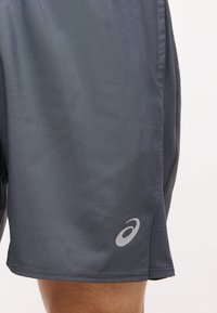 Σκούρο γκρι αθλητικό σορτς με αντανακλαστικό λογότυπο Asics κοντά στο κάτω τελείωμα, πάνω στο μηρό ενός ατόμου.