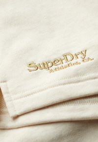 Tissu crème avec une texture douce, arborant un fil doré en relief avec "SuperDry" et "Athletic, CA" dans le coin inférieur droit.