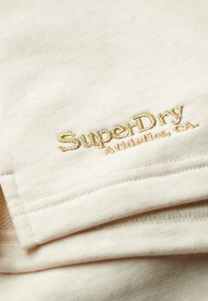 Roomkleurige stof met een zachte textuur, voorzien van reliëf gouden stiksel van "SuperDry" en "Athletic, CA" in de rechterbenedenhoek.