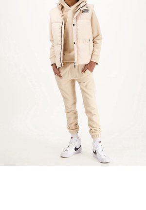 Person wearing beige hoodie, matching jogger pants, cream-colored puffer vest, and white Nike high-top sneakers, standing with hands in pockets.