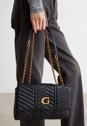 Ręka trzyma czarną pikowaną skórzaną torebkę z złotymi łańcuszkowymi paskami i złotym logo „G”, noszoną z ciemnymi dżinsami i szarą bluzką.