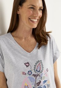 Leende kvinna med långt brunt hår bär en ljusgrå V-ringad tröja med färgglad blommig print på framsidan, stående mot en enfärgad bakgrund.