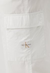Biele cargo šortky z ľahkej tkaniny so štvorecovou náprsnou vreckom, s logom "Calvin Klein Jeans" v zlatej a čiernej farbe.