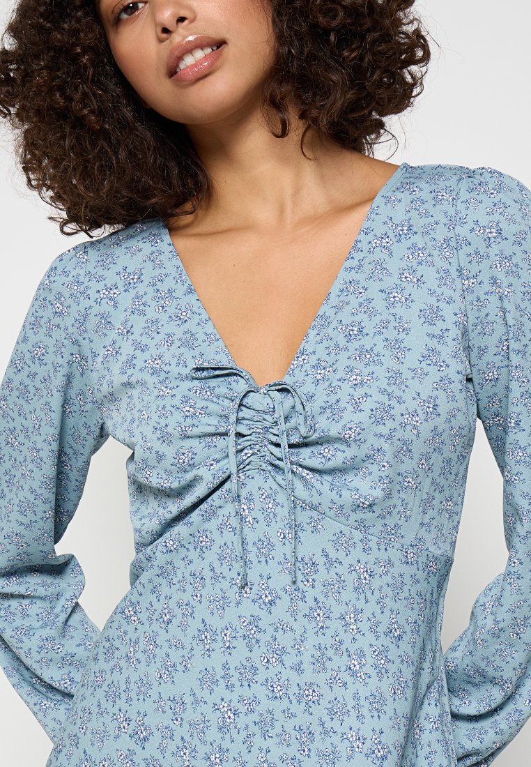 Vestido floral azul claro con un escote en V profundo, detalles fruncidos en el busto y mangas largas con un tejido texturizado y pequeños acentos blancos.