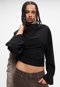 Top negro cropped de manga larga con mangas sueltas y voluminosas y un cuello alto, combinado con pantalones marrones de cintura alta, que exhiben una textura suave.