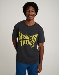 Σκούρο γκρι βαμβακερό t-shirt με κίτρινο γραφικό "Stranger Things" σε στυλ φλόγας στο μπροστινό μέρος, κοντά μανίκια και άνετη εφαρμογή.