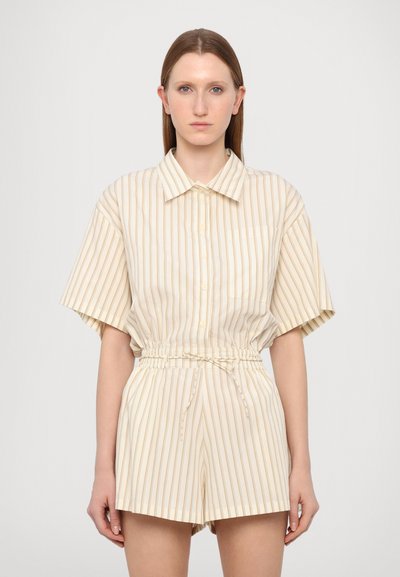 Completo composto da una camicia a maniche corte e pantaloni corti di colore crema, con sottili righe verticali in marrone chiaro, vita arricciata e chiusura con bottoni sul davanti.