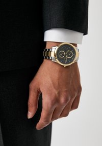 Zlato in srebro nerjaveče jekleno uro na zapestju, ki ima črn premaz, dve subdiali in teksturiran kovinski pas. Eleganten in sodoben dizajn.