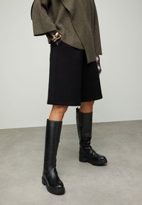 Zwarte knielange laarzen met grove zolen, gemaakt van leer. Gecombineerd met een oversized olijfgroene trui en zwarte shorts. Gladde textuur.