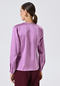 Donna con codino che indossa una blusa lunga a maniche corte in seta lavanda e pantaloni bordeaux, vista di spalle su uno sfondo neutro.