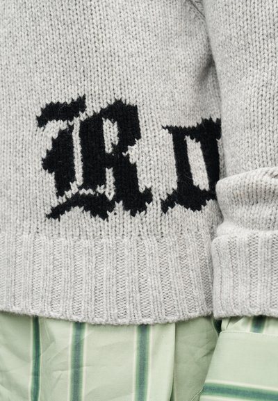 Ανοιχτό γκρι πλεκτό πουλόβερ με μαύρο γοτθικό κείμενο πάνω σε μανίκι και μανσέτα πράσινου ριγέ πουκαμίσου ορατά στο κάτω μέρος.