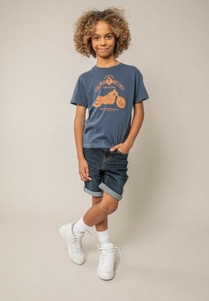 Enfant aux cheveux bouclés portant un t-shirt bleu avec un motif de moto orange, un short en jean foncé retroussé, des chaussettes blanches et des baskets blanches.