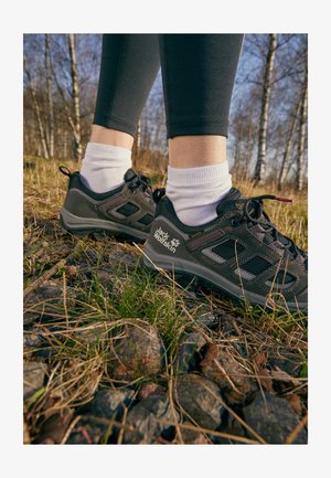 Zwarte wandelschoenen met een mesh bovenkant, paarse accenten en een robuuste zool, gedragen met witte sokken, op een achtergrond van gras en stenen.