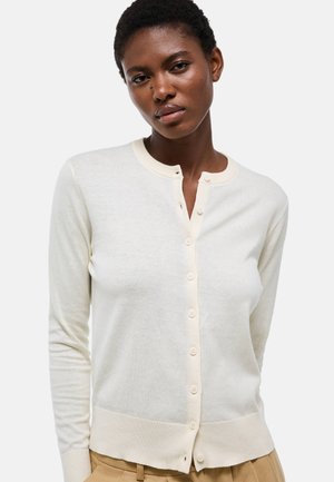 Jeune femme portant un cardigan crème boutonné et un pantalon beige, faisant face avec une expression neutre sur un fond uni.