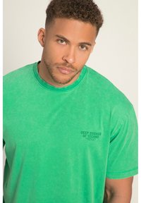 Camiseta de algodón verde, de cuello redondo, con una pequeña impresión de texto "SONIDOS PROFUNDOS DEL TECHNO" en el lado izquierdo del pecho. Acabado suave y texturizado.