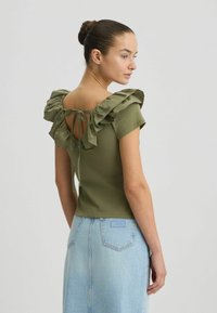Olivengrøn kortærmet top med flæsekrave og bindebånd bagpå, parret med en lyseblå denimnederdel med en baglomme.