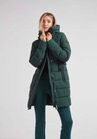 Chaqueta de plumas verde oscura con cuello alto, acolchado horizontal, cremallera frontal y botones a presión. Presenta un parche de logo en el hombro.