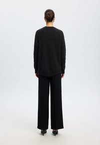 Pulover negru, pufos, cu mâneci lungi, gât rotund și spate puțin mai lung, asortat cu pantaloni negri cu picior larg și tocuri negre elegante.