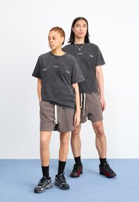 To modeller har på seg mørkegrå, oversized t-skjorter med subtile trykkdetaljer, kombinert med brune shorts med snor i livet og svarte joggesko.