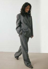 Siva volnena mešanica suit, ki vključuje dvoredni blejzer z razrezanimi reverji in široke hlače. Teksturo tkanine dopolnjujejo subtilni vzorci.