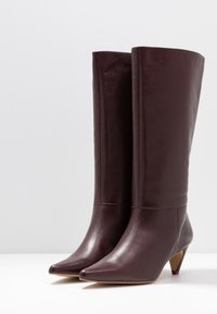 Bottes en cuir bordeaux montant jusqu'au genou, avec un bout pointu et un talon de hauteur moyenne, présentant une texture lisse et un design minimaliste.