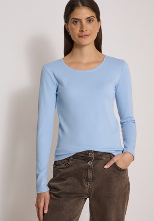 Donna con capelli lunghi e scuri che indossa una camicia azzurra a maniche lunghe e pantaloni marroni, con una mano in tasca, su uno sfondo semplice.