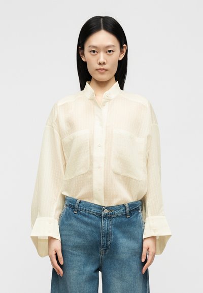 Jeune femme aux cheveux noirs et raides portant un chemisier ample beige clair boutonné et un jean bleu taille haute sur un fond uni.