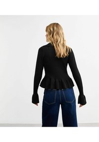 Czarny sweter z prążkowanej dzianiny z peplum, z długimi rękawami i rozkloszowanymi mankietami. Noszony z niebieskimi dżinsami o wysokim stanie.