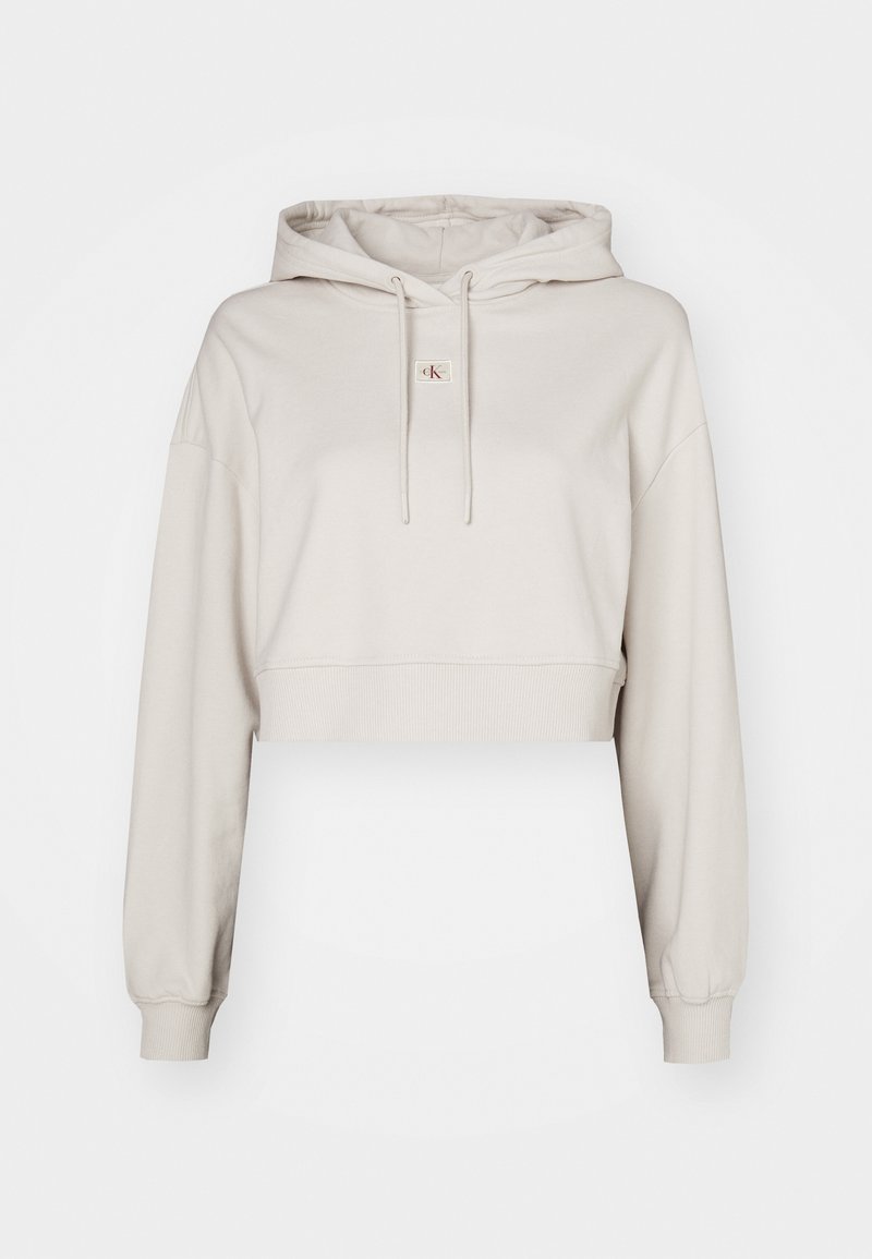 Beige Crop-Hoodie aus weichem Stoff, mit einer Kordelzugkapuze, langen Ärmeln und einem kleinen Logopatch auf der Brust.
