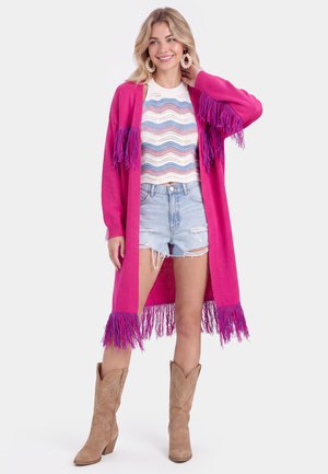Smilende kvinde iført en lyserød cardigan med frynser, en stribet strik-top, revnede denimshorts, store øreringe og brune cowboystøvler, stående med hånden nær håret.