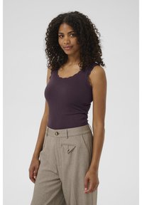 Débardeur violet foncé à côte avec décolleté festonné, associé à un pantalon beige à motifs avec une poche triangulaire et un bouton doré.