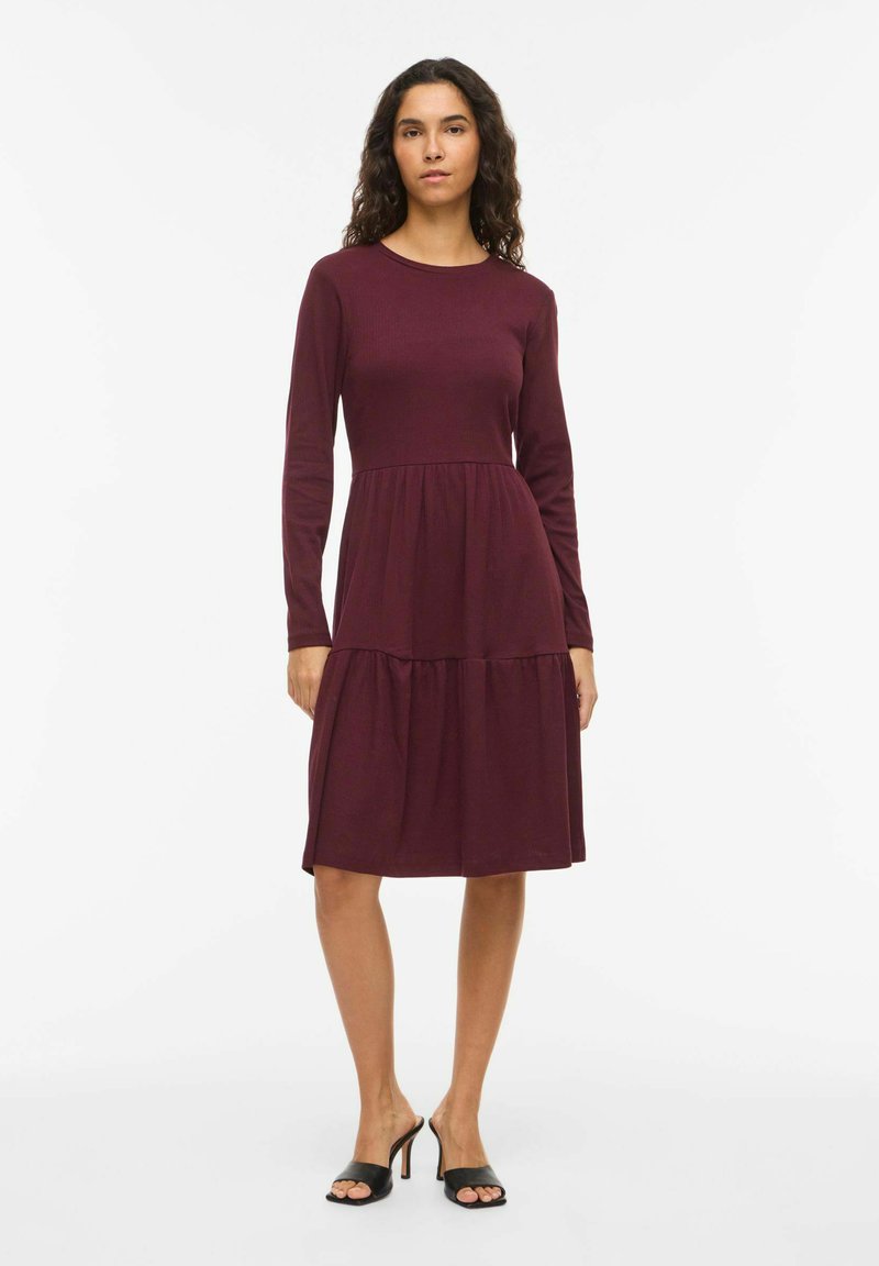 Robe bourgogne à manches longues avec une jupe à volants, réalisée en tissu doux. Elle présente un col rond et un corsage ajusté pour une coupe décontractée.