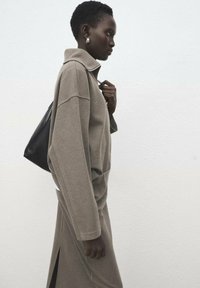 Prevelik bež pulover iz flisa z visokim ovratnikom, kombiniran z usklajeno krilo s stranskim razporkom. Črna torba za na ramo.