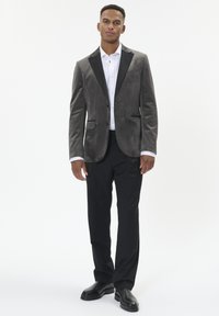 Grå ruskindsjakke med sorte klapper, parret med en hvid skjorte og sorte bukser. Har en struktureret pasform og subtile lomme detaljer.