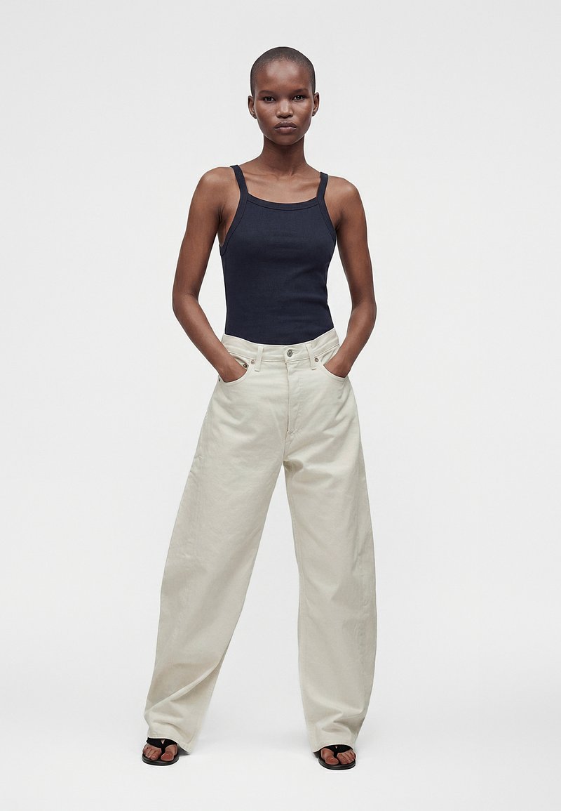 Femme portant un haut noir sans manches, un pantalon large crème et des sandales noires, debout les mains dans les poches devant un fond blanc uni.