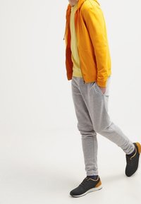 Sudadera con capucha amarilla con cremallera, camiseta verde, pantalones deportivos grises y zapatillas negras con detalles naranjas. La textura es suave con un ajuste informal.