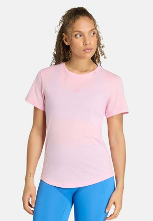 Femme aux cheveux bouclés portant un t-shirt rose clair à manches courtes et un pantalon bleu vif, regardant vers sa gauche sur un fond blanc uni.